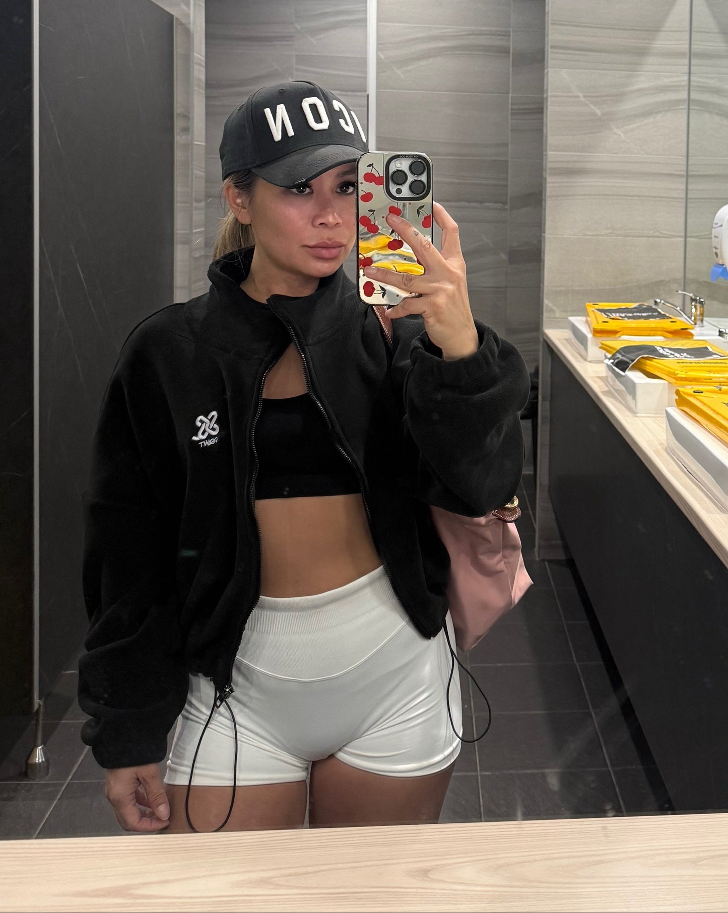 Woman wearing a black
 colour fleece cropped zip-up jacket with high collar and embroidered logo, paired with white shorts, taking selfie at the gym bathroom