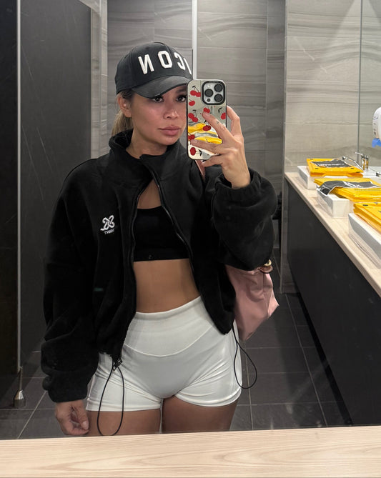 Woman wearing a black
 colour fleece cropped zip-up jacket with high collar and embroidered logo, paired with white shorts, taking selfie at the gym bathroom