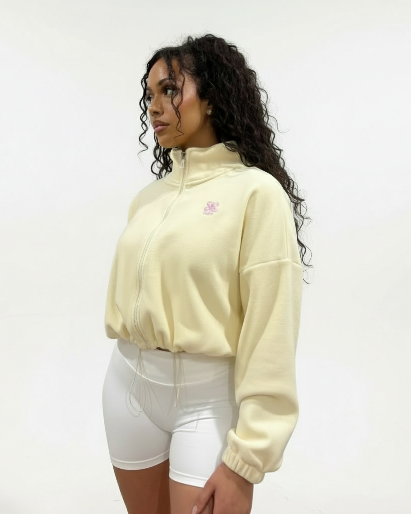 Woman wearing a buttermilk colour fleece cropped zip-up jacket with high collar and embroidered logo, paired with white shorts, standing against a plain studio background.