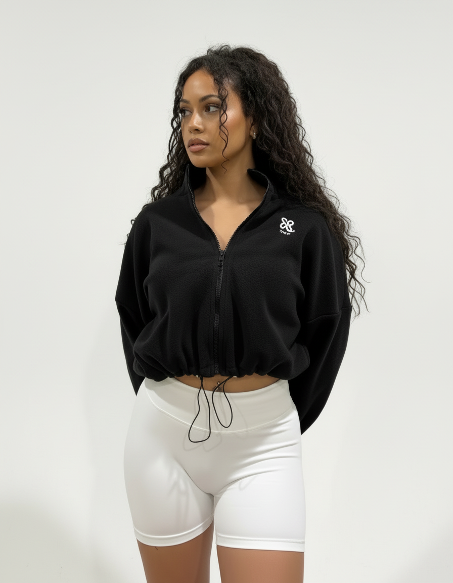 Woman wearing a black
 colour fleece cropped zip-up jacket with high collar and embroidered logo, paired with white shorts, standing against a plain studio background.