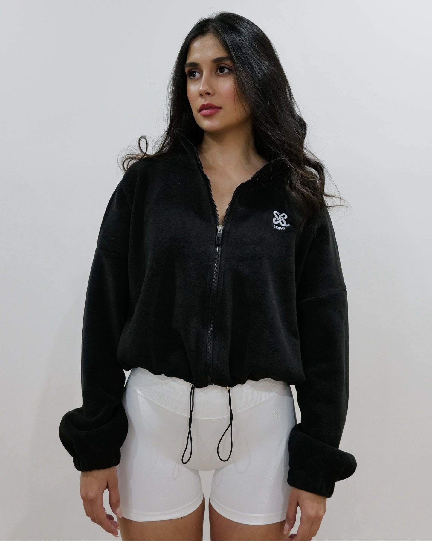 Woman wearing a black
 colour fleece cropped zip-up jacket with high collar and embroidered logo, paired with white shorts, standing against a plain studio background.