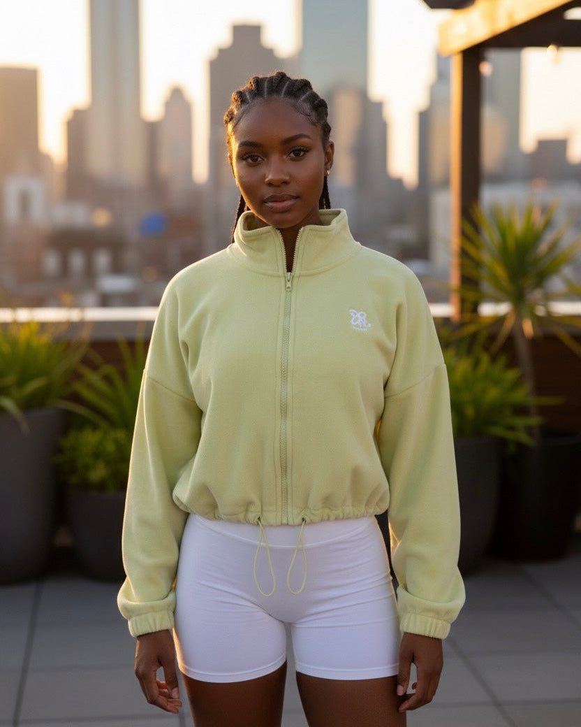 Woman wearing a light green colour fleece cropped zip-up jacket with high collar and embroidered logo, paired with white shorts, standing against a building in the city 