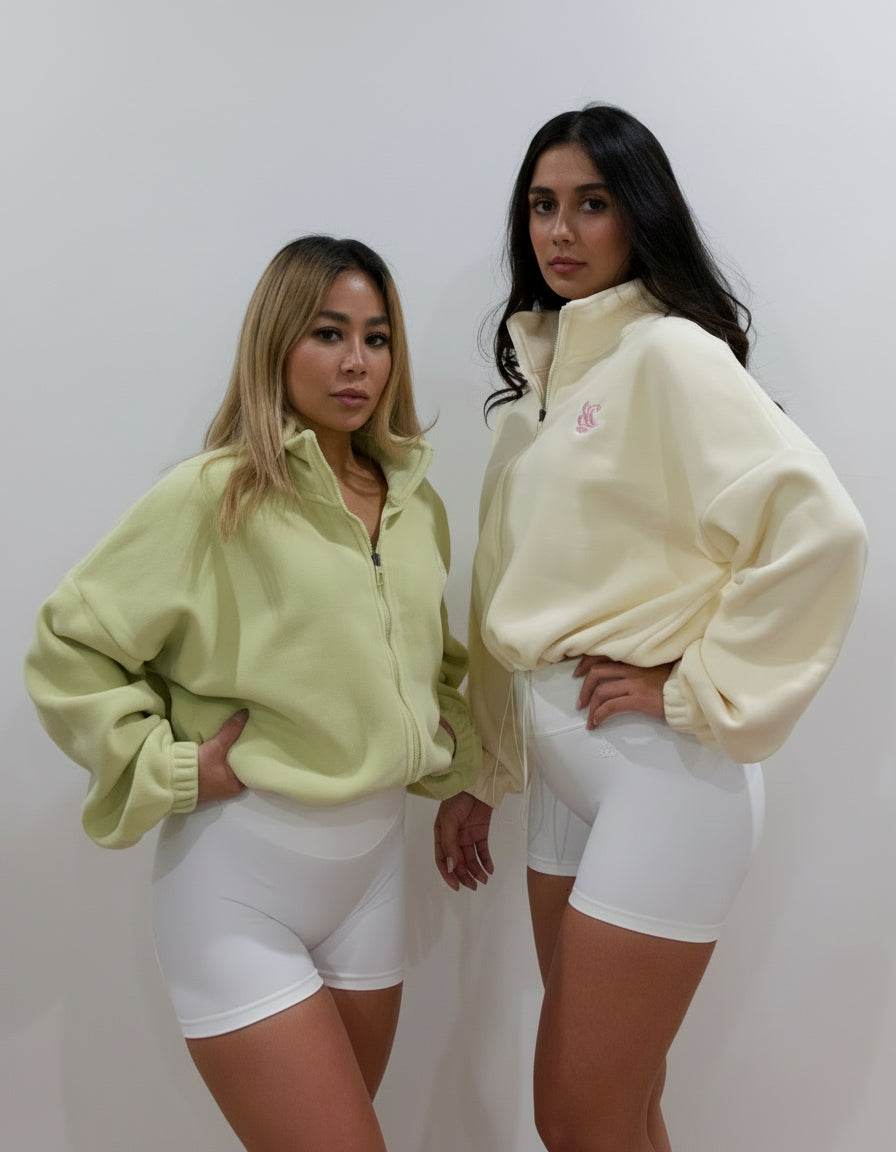 Two women wearing a buttermilk colour and light green colour fleece cropped zip-up jacket with high collar and embroidered logo, paired with white shorts, standing against a plain studio background.