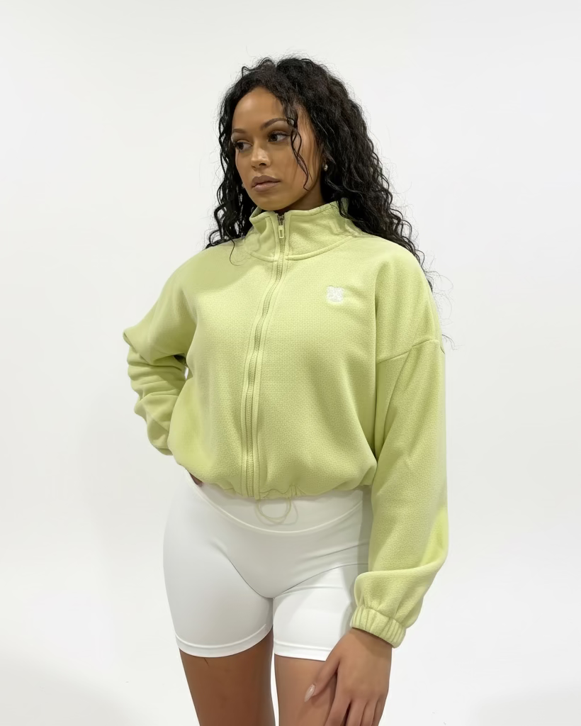 Woman wearing a light green colour fleece cropped zip-up jacket with high collar and embroidered logo, paired with white shorts, standing against a plain studio background.