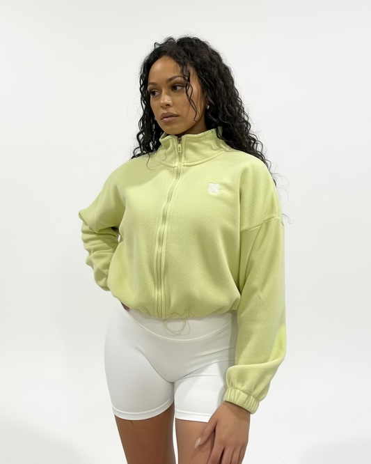 Woman wearing a light green colour fleece cropped zip-up jacket with high collar and embroidered logo, paired with white shorts, standing against a plain studio background.
