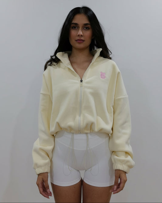Woman wearing a buttermilk colour fleece cropped zip-up jacket with high collar and embroidered logo, paired with white shorts, standing against a plain studio background.